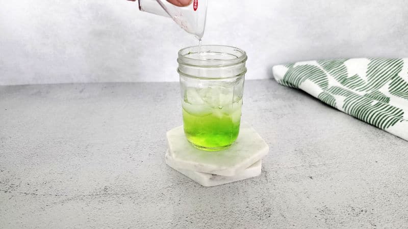 Clear liquid pouring into a small mason jar with green liqueur in it for a Scooby Snack Shot