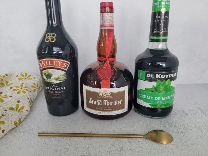 Baileys Irish Cream, Grand Marnier, and Creme de menthe bottles lined up next to a gold bartending spoon and cloth napkin 