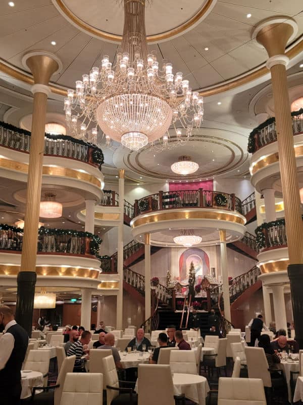 Large crystal chandelier hanging over white tables and chairs with multiple staircases in the background