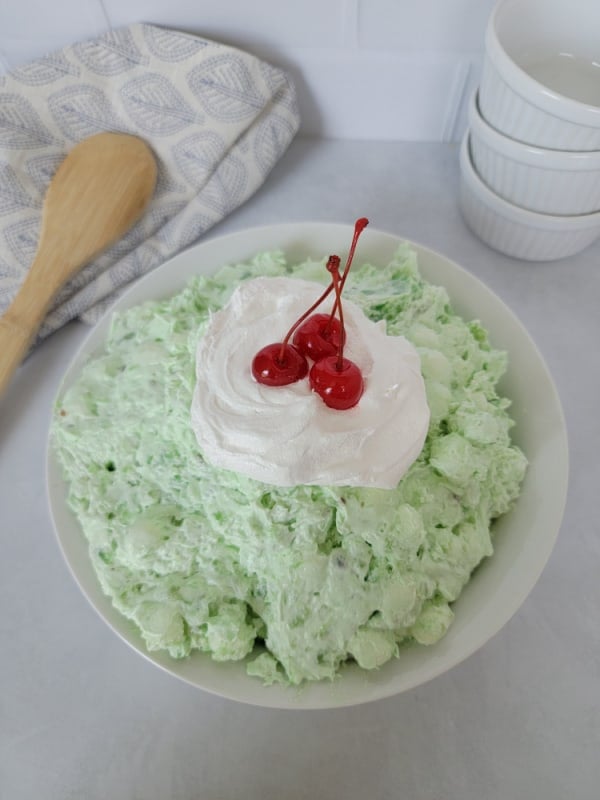 Classic Watergate Salad in a white bowl with a dollop of Cool Whip and Maraschino Cherries next to a wooden spoon and three white bowls