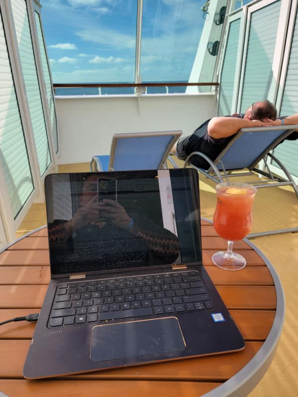 Tammilee taking a photo of a laptop next to a tropical drink with john lounging on a lounge chair with the ocean in the distance