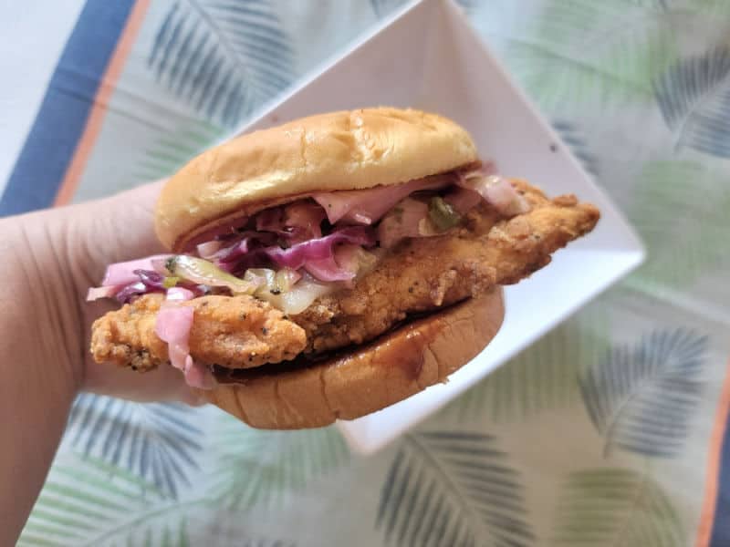 Hand holding a chicken sandwich with slaw over a white bowl and tropical napkin