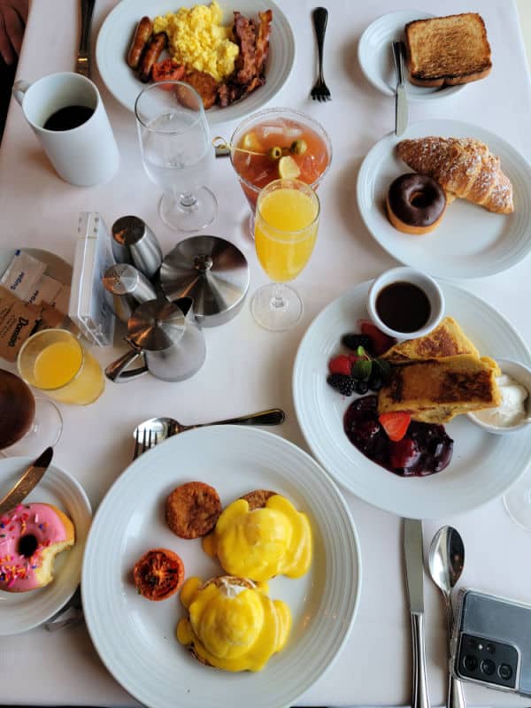 breakfast spread over a white table cloth including eggs benedict, French toast, a donut, croissant, Bloody Mary and Orange Juice