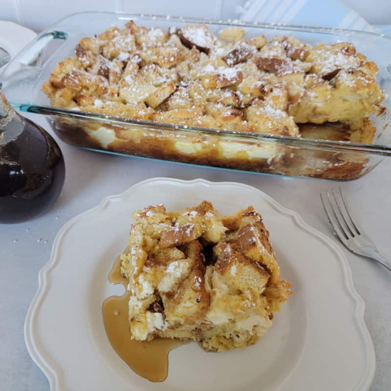 square of overnight French toast on a white plate in front of a casserole dish filled with French Toast casserole