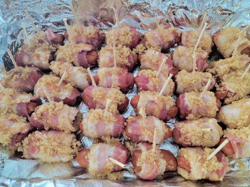Rows of unbaked bacon wrapped lit'l smokies in an aluminum foil lined baking dish
