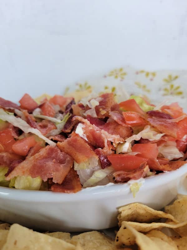 Bacon, diced tomato, and lettuce in a white bowl next to tortilla chips and a cloth napkin 