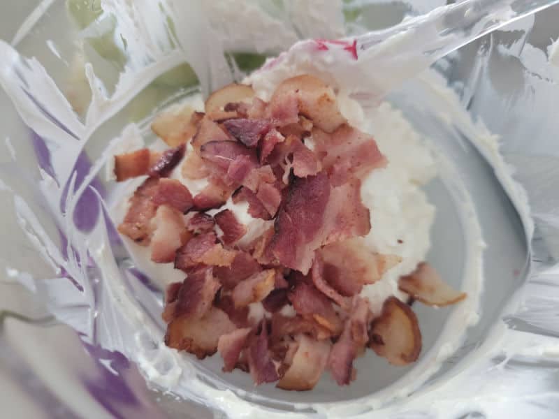 Bacon pieces on top of the cream cheese mixture in a glass bowl with a spatula
