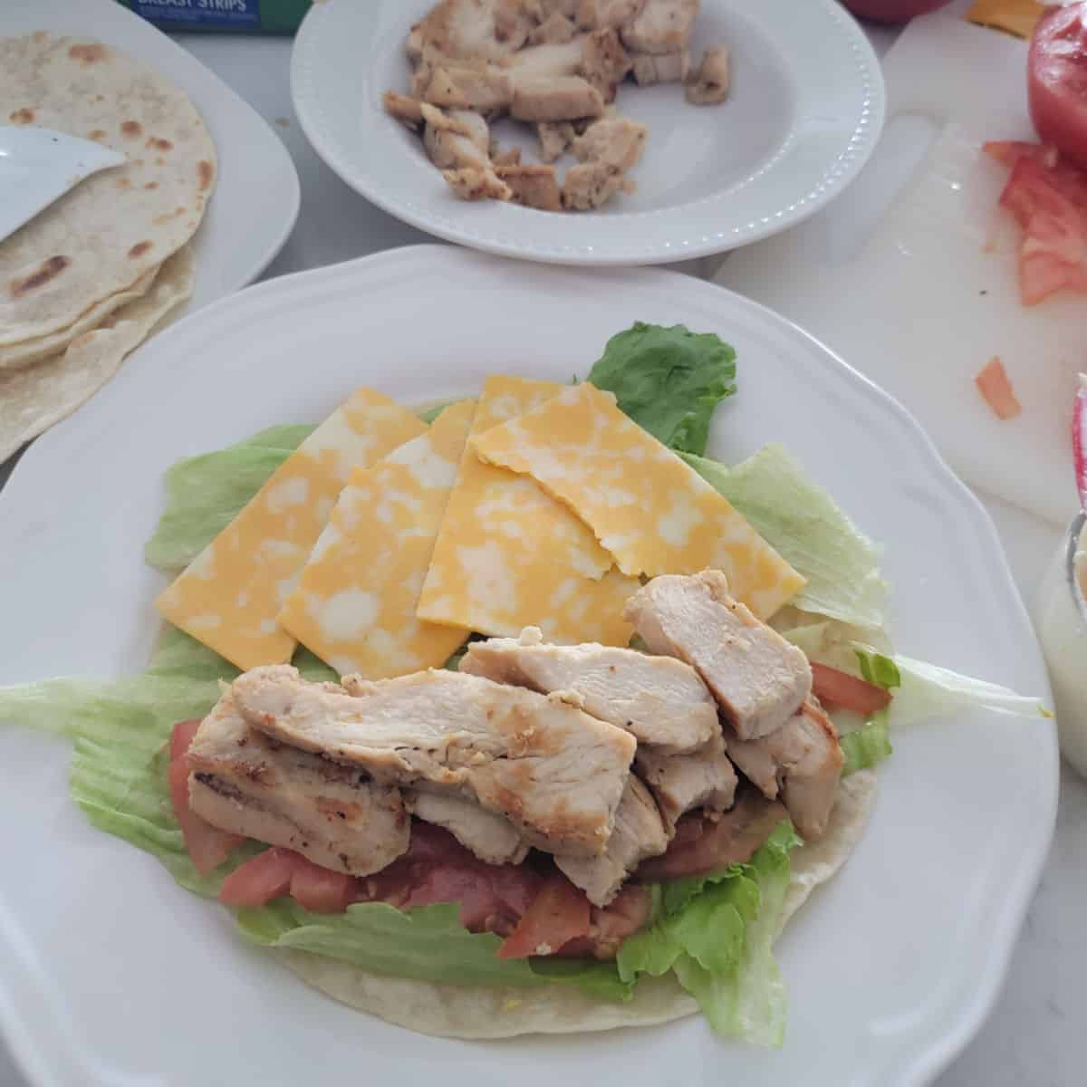 chicken wrap with lettuce and cheese on a white plate with a plate of chicken in the background