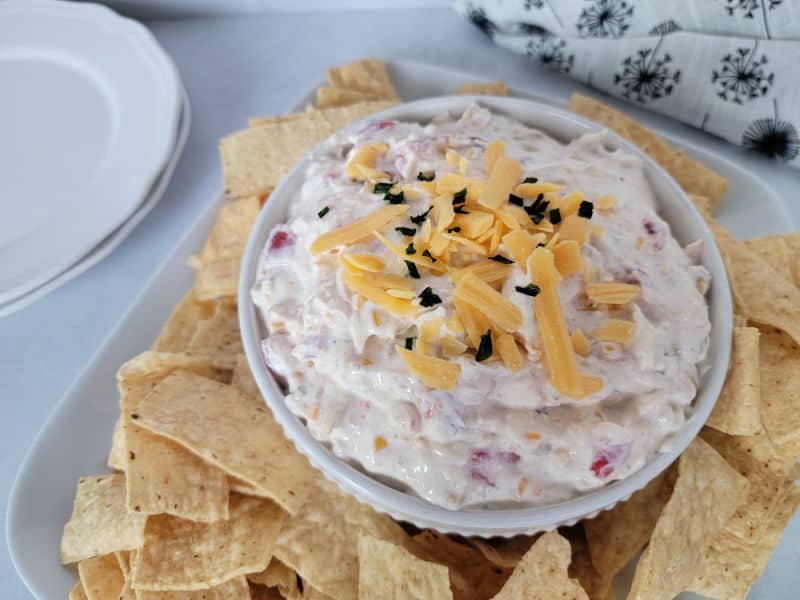 Fiesta dip garnished in shredded cheese in a white bowl surrounded by tortilla chips on a white platter next to a stack of plates