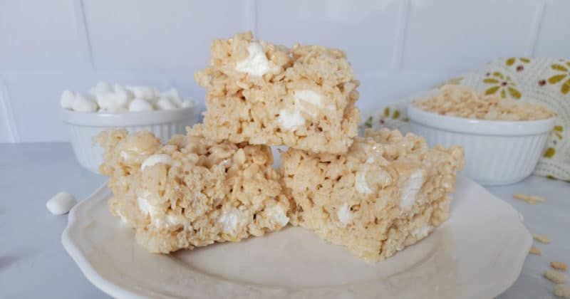 Stack of Rice Krispie Treats on a white plate with marshmallows and rice krispie cereal spread out