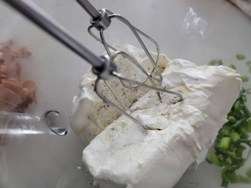 Mixer beaters in a glass bowl with cream cheese, ranch dressing mix, and green onions