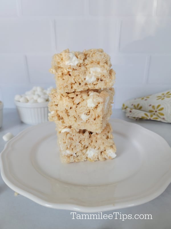 Stack of 3 Rice Krispie Treats on a white plate with mini marshmallows in a bowl behind 