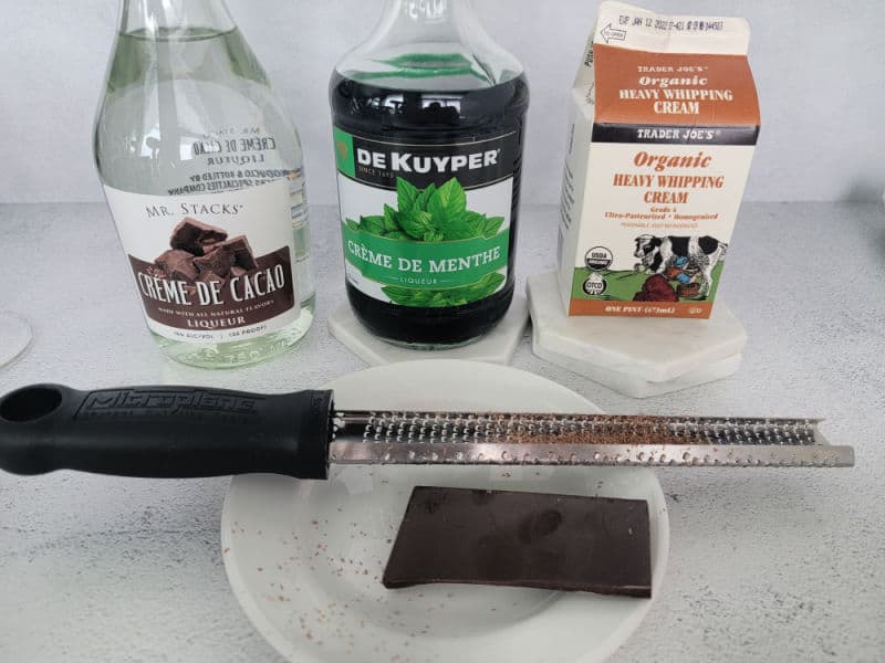 Creme De Cacao, Creme de Menthe, Heavy Whipping Cream bottles behind a plate with chocolate on it and a zester