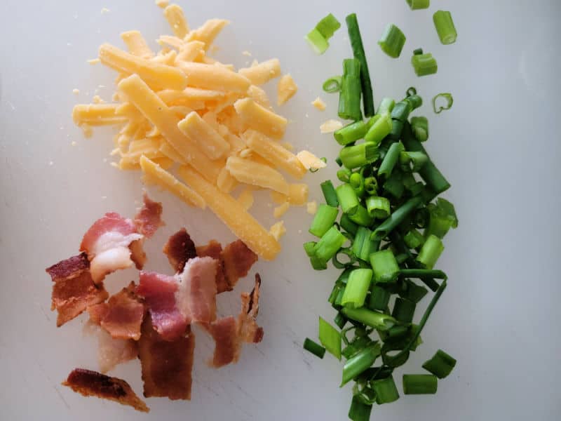 shredded cheddar cheese, bacon pieces, and chopped green onion on a cutting board. 