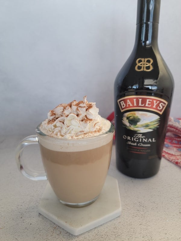 Baileys Coffee with whipped cream garnish in a glass coffee mug next to a bottle of Baileys