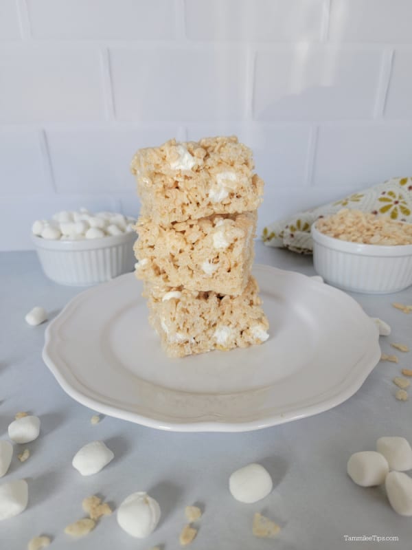 Stack of three Rice Krispie Treats on a white plate with mini marshmallows and rice cereal surrounding the plate