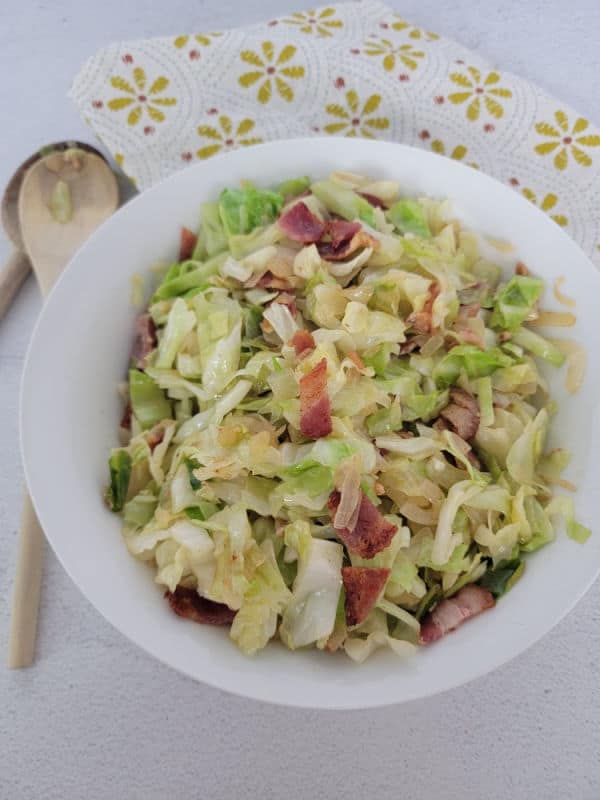 Bacon and shredded cabbage in a white bowl next to a wooden spoon and cloth napkin