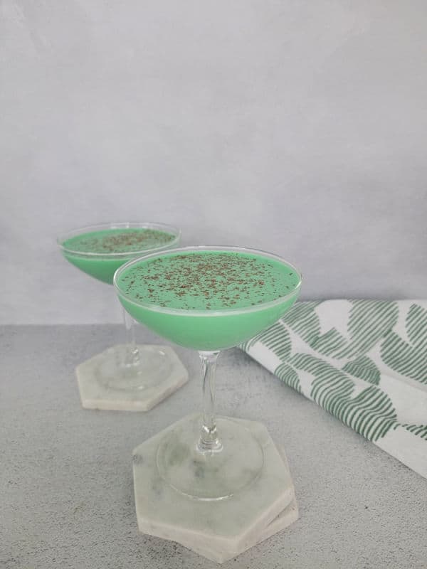 Two green cocktails in coupe glasses on white coasters by a linen towel 
