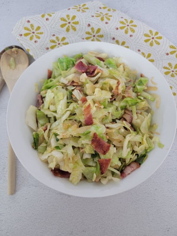 Cabbage and bacon in a white bowl next to wooden spoons and a cloth napkin