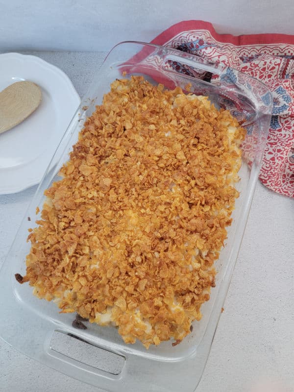 Baked funeral potatoes in a glass baking dish next to a white plate and wooden spoon. 