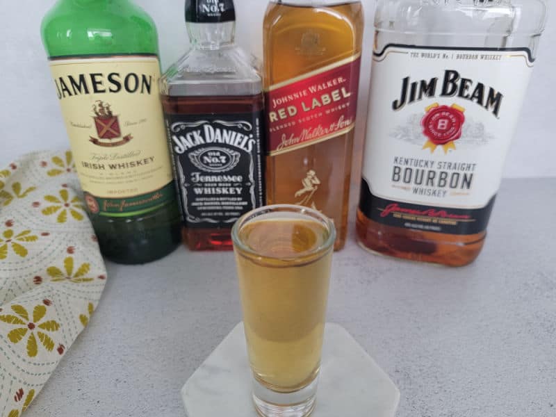 Four horsemen shot in a shot glass on a white coaster in front of bottles of Jameson Irish Whiskey, Jack Daniels, Johnnie Walker and Jim Beam