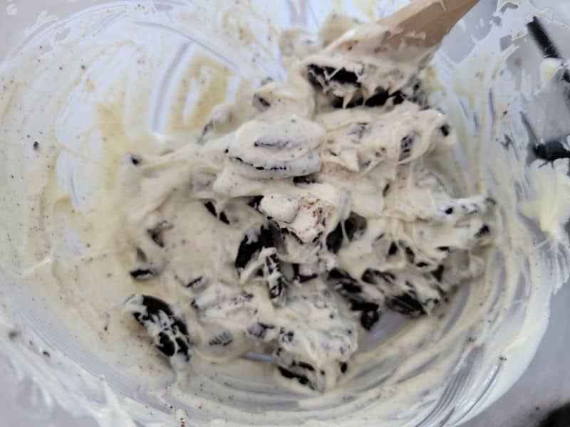 Oreo pieces mixing into melted white chocolate in a glass bowl with a wooden spoon 