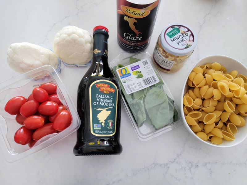 tomatoes, mozzarella balls, olive oil, balsamic glaze, basil, minced garlic, and pasta shells in a bowl on a white counter 