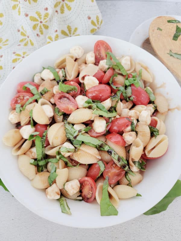 Caprese Pasta salad in a white bowl next to a wooden spoon and cloth napkin