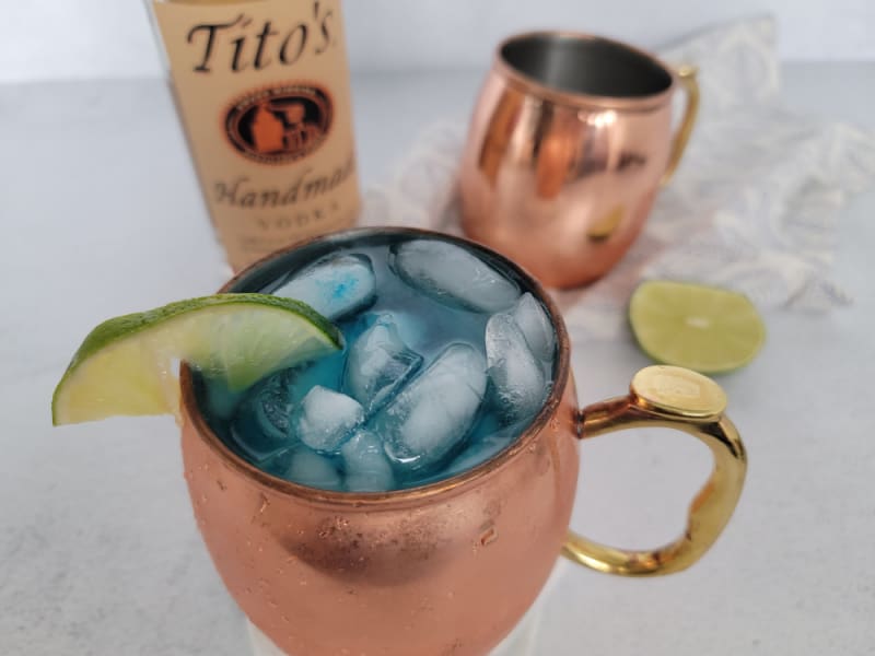 Blue Moscow Mule in a copper mug next to a bottle of Titos Vodka and a lime wheel