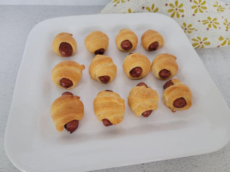 Baked pigs in a blanket lined up on a white platter next to a cloth napkin