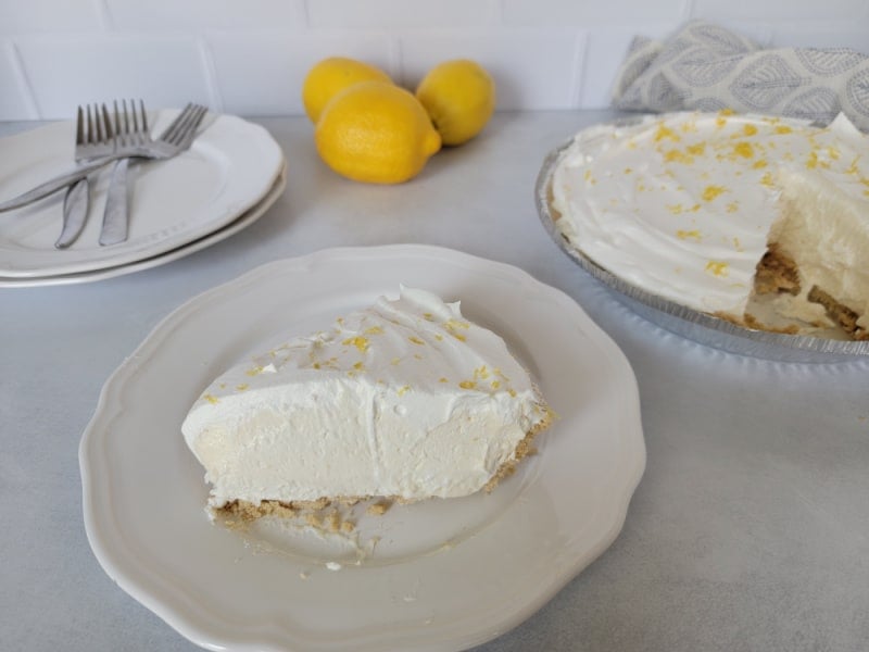 Slice of no bake lemon pie on a white plate with the full pie in the background. 