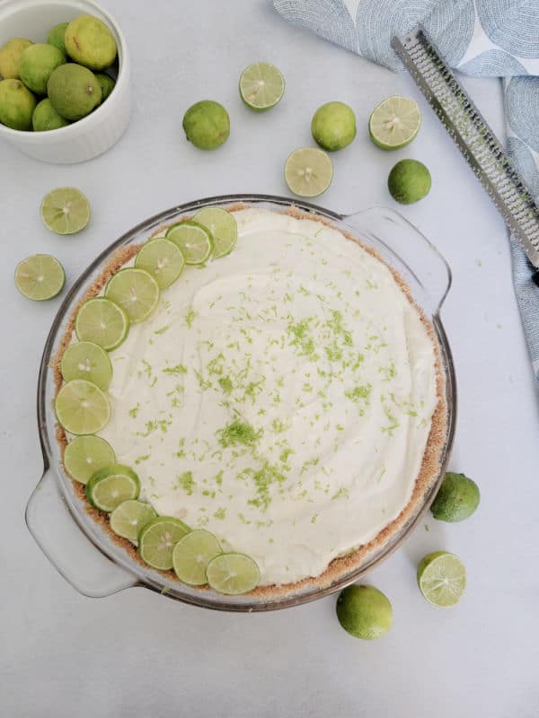 Bowl of key limes next to a zester and glass pie dish with key lime pie in it