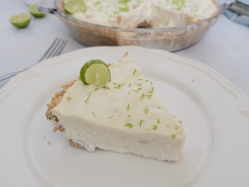 Slice of no bake key lime pie on a white plate next to a fork with the pie in the background