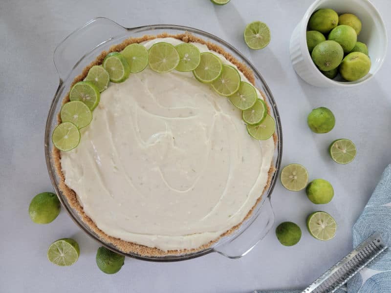 Key lime circles spread around the edge of a no bake key lime pie in a glass baking dish next to a bowl of limes