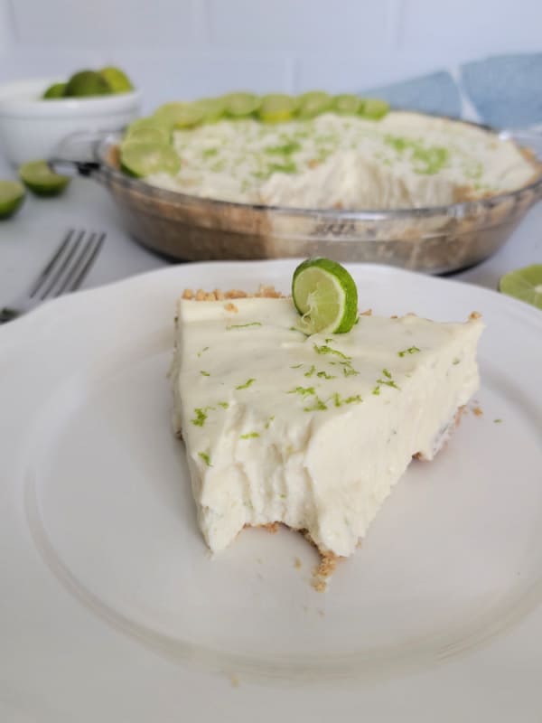 Slice of key lime pie with a bit taken of the front on a white plate next to a fork and the pie in the background. 