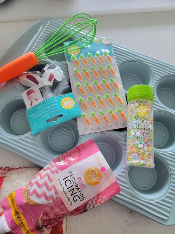 cupcake pan, bunny ears, carrot whisk, carrot candy, sprinkles, and pink icing on a white counter. 