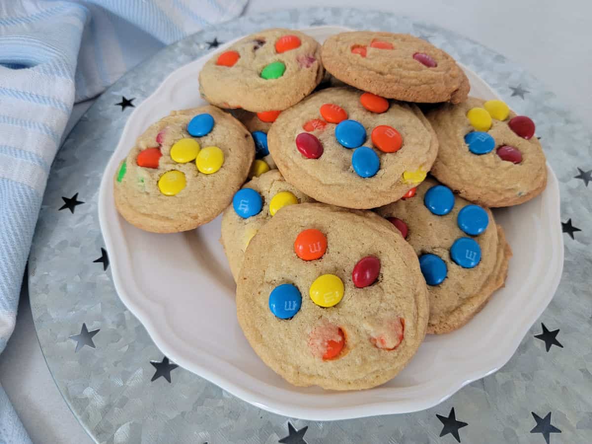 Colorful M&M Cookies on a white plate on a silver platter next to a cloth napkin