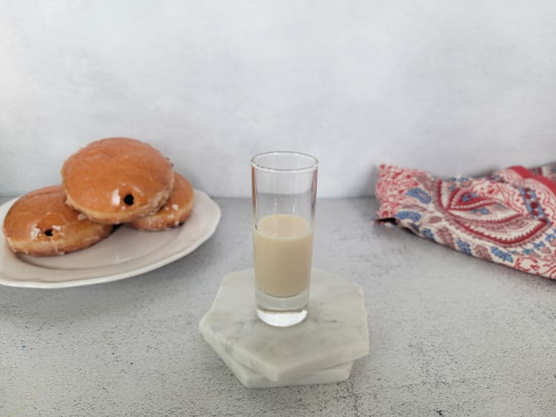 Plate of jelly doughnuts next to a shot glass half full with cream liquid sitting on white coasters. 