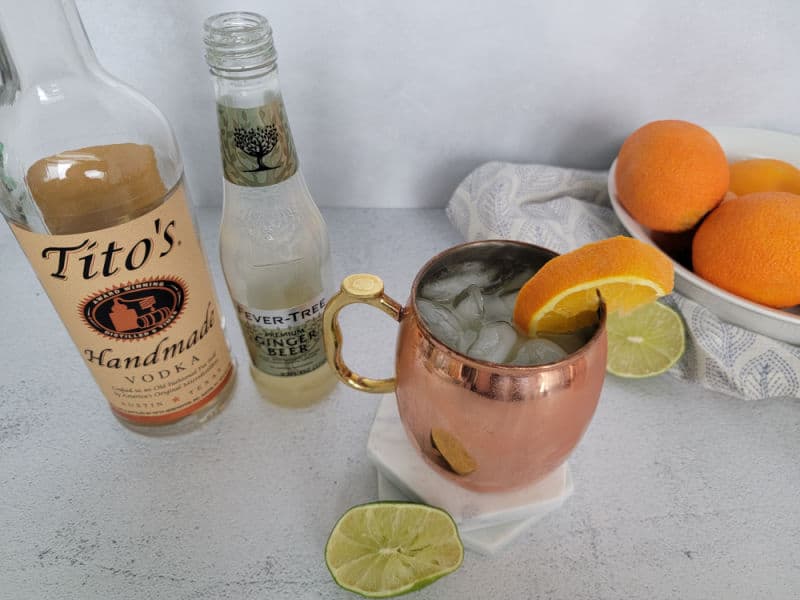 Orange Moscow mule in a copper mug next to a bowl of orange, bottle of Tito's vodka, and ginger beer