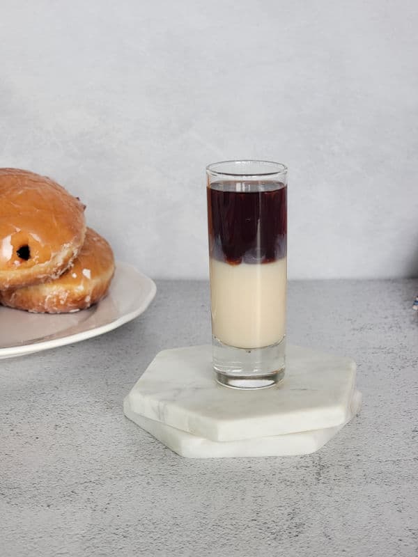 layered purple and cream shot on two white coasters next to a white plate with jelly doughnuts. 