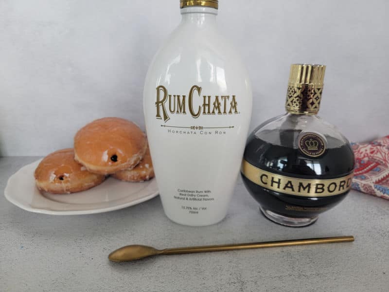 Jelly doughnuts on a white plate next to a bottle of RumChata and Chambord and a gold bar spoon