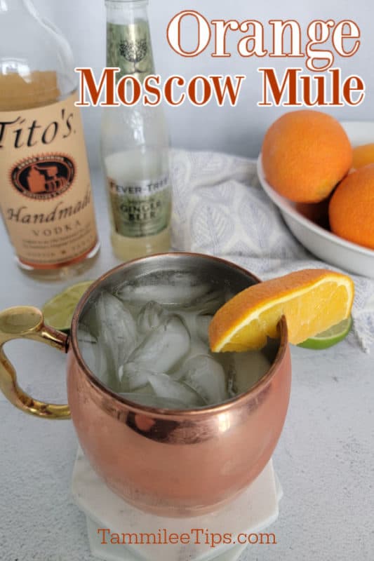 Orange Moscow Mule text over a bottle of titos vodka, ginger beer, a bowl of oranges, and an orange moscow mule in a copper mug