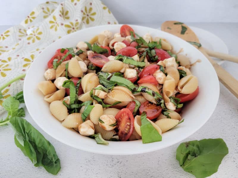 Caprese pasta salad in a white bowl with basil leaves and a wooden spoon next to it. 