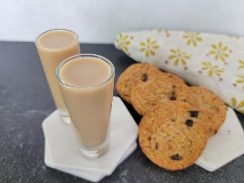 Two cocktail shots on a white coaster next to a stack of oatmeal cookies and a cloth napkin