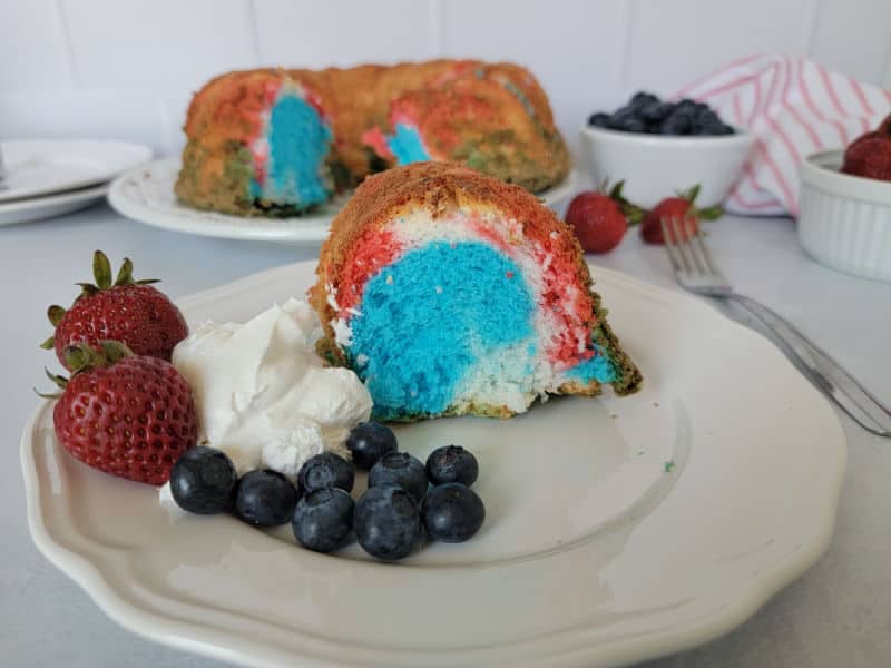 Red white and blue angel food cake slice on a white plate with whipped cream, strawberries, and blueberries next to the full cake. 