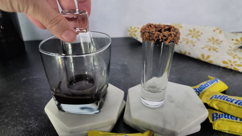 Shot glass dipping into chocolate syrup in a glass next to a shot glass on a coaster