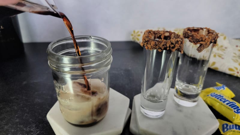 Brown liquid pouring into a small mason jar on a white coaster next to to shot glasses rimmed in chocolate and Butterfinger pieces