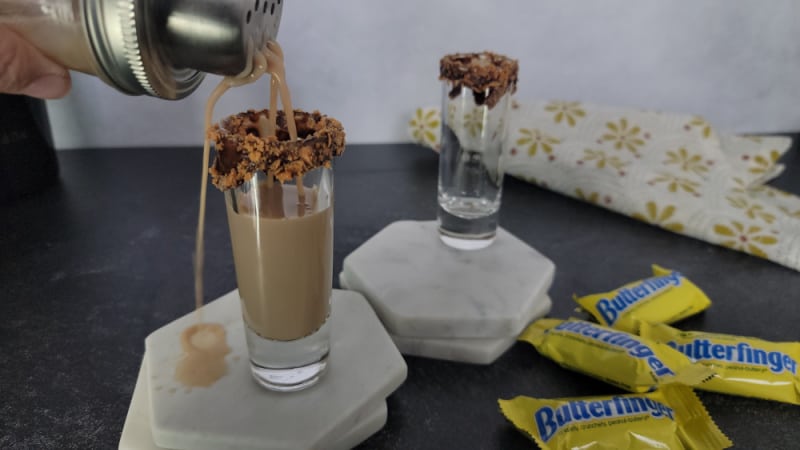 Creamy liquid pouring into a shot glass rimmed with chocolate and butterfinger pieces