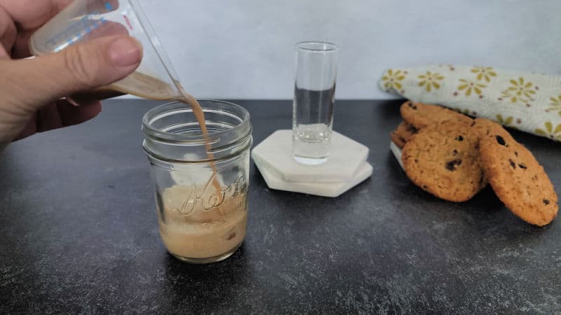 Hand pouring liquid into a mason jar next to a shot glass and oatmeal cookies