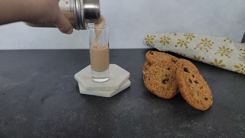 Cocktail shaker pouring into a shot glass next to oatmeal cookies and a cloth napkin